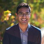 A man wearing glasses and a black suit jacket stands and smiles