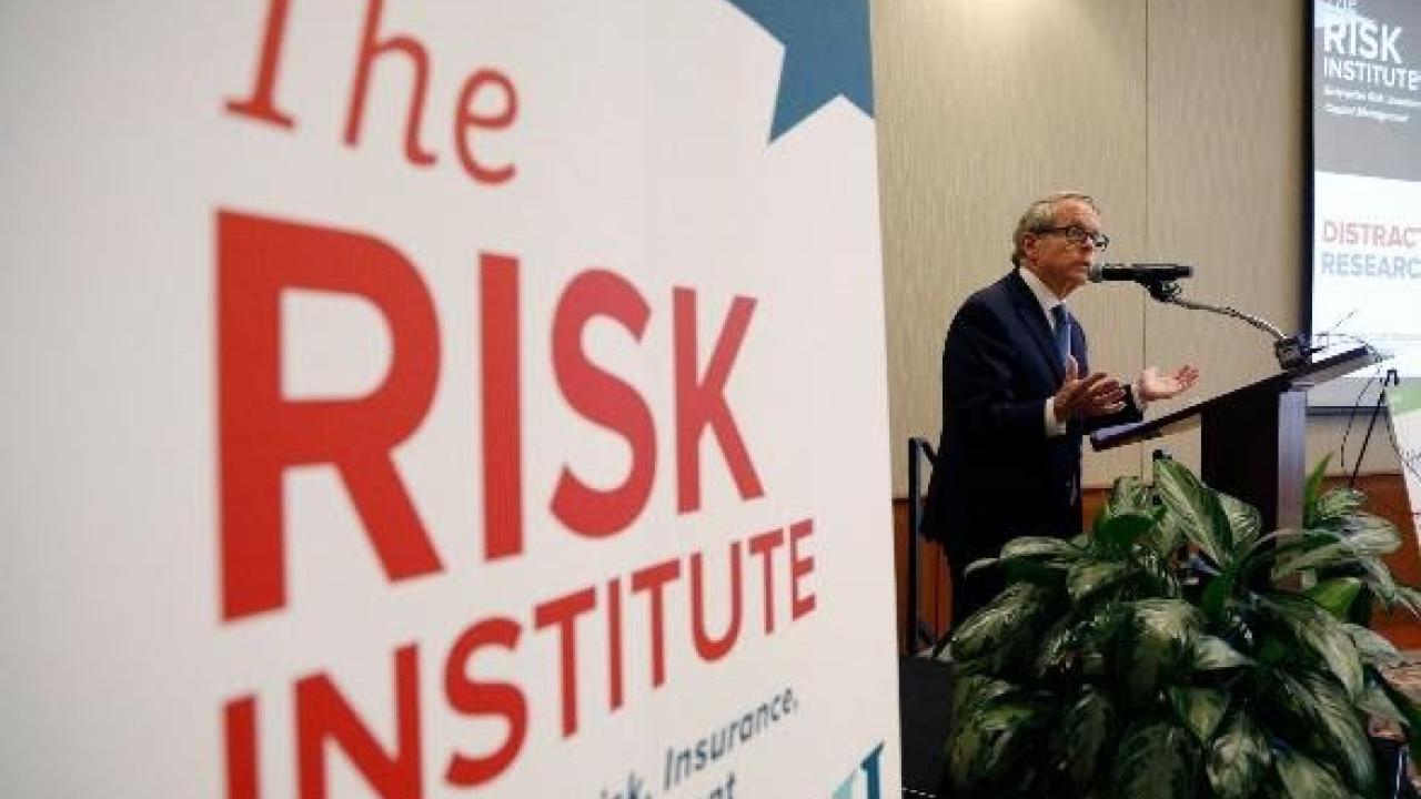 Presenter speaking with a sign that reads "The Risk Institute" in the foreground.